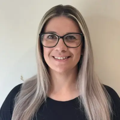 Smiling woman with long blonde hair and glasses against a plain wall.